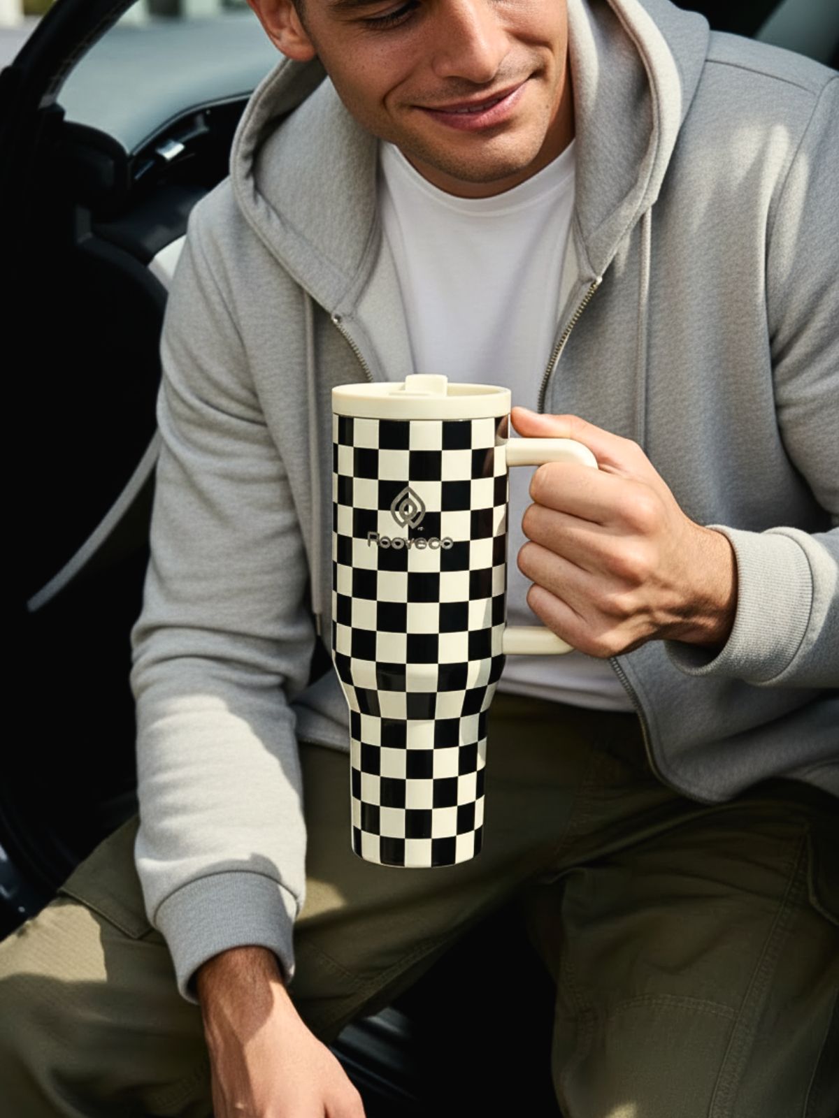 Man sitting by a car holding a checkered Rooveco tumbler. Casual modern lifestyle product shot.